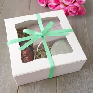 Peut inclure: Boîte cadeau blanche avec une fenêtre transparente, remplie d'une petite bouteille de liquide, d'un petit pot et de papier déchiqueté. La boîte est attachée avec un ruban vert.