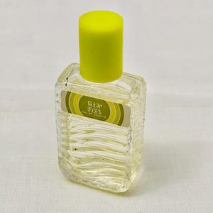 Op de afbeelding: Een kleine, heldere glazen parfumfles met een textuurontwerp. De fles heeft een felgele dop en een etiket met de tekst "Gin Fizz Eau de Toilette" in groen en geel. De fles is gevuld met een heldere vloeistof.