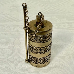 May include: A vintage brass cylindrical container with a decorative chain and a long, thin handle. The body features intricate openwork designs. The container is likely used for storing small items or as a decorative piece.