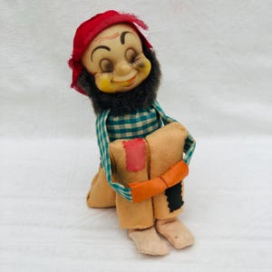 Puede incluir: Una muñeca vintage con una cara sonriente, ojos cerrados y una gorra roja. La muñeca tiene barba negra, una camisa a cuadros azul y blanca y pantalones color canela con parches. Una banda naranja envuelve las piernas de la muñeca.