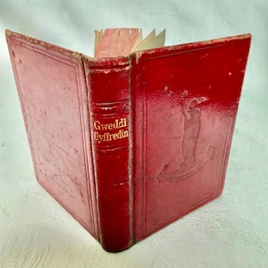 Puede incluir: Un libro antiguo de tapa dura roja con el título Gweddi Gyffredin en el lomo. La portada presenta una figura en relieve y un diseño de estandarte. El libro está ligeramente abierto, mostrando páginas claras en el borde superior.