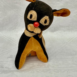 May include: Vintage plush toy dog with black and gold coloring. The dog has large, round eyes, a red nose, and a tan scarf. The ears are brown. The toy is sitting upright.
