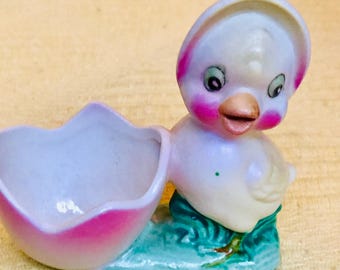 May include: A white ceramic chick figurine with pink cheeks and a blue and green base. The chick is holding a pink and white egg shaped dish.