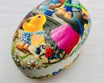 May include: A colorful Easter egg-shaped tin with a whimsical scene of a chick, a mouse, and a ladybug. The tin features a white background with a scalloped edge.