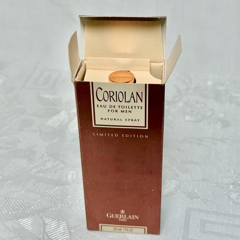 May include: An open box of Guerlain Coriolan Eau de Toilette for Men. The brown box has the product name and details in white text. The bottle is visible inside the box, with a gold-colored cap. The box is a limited edition.