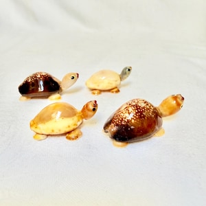 May include: Four small turtle figurines crafted from polished seashells. Two turtles feature dark brown shells with white speckled patterns, while the other two have lighter tan and cream-colored shells. Each has a smooth, rounded shell.