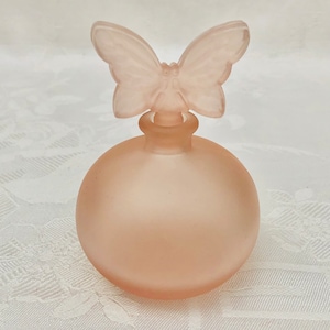 May include: A frosted glass perfume bottle with a butterfly-shaped stopper. The bottle is a pale pink color and has a round shape.