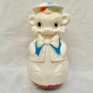 May include: Vintage white bear-shaped toy with a sailor hat and bow tie. The hat has blue, orange, and green accents. The bear wears a blue vest and a plaid jacket. The bottom is red.
