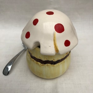 Peut inclure: Un pot à sucre en céramique blanche en forme de champignon avec des pois rouges et une base jaune. Le couvercle est ouvert et une cuillère en argent se trouve à côté du bol.