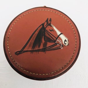 May include: A brown leather coaster with a white stitched border and a painted image of a brown horse's head with a white bridle.