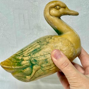 May include: A vintage duck figurine in shades of yellow and green. The duck has detailed feather patterns and a curved neck. The figurine is held by a hand, showcasing its size and texture. The duck's beak is a darker shade of brown.