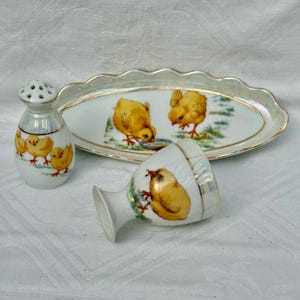 May include: A vintage porcelain Easter-themed set featuring a salt shaker, an egg cup, and a serving dish. The set is decorated with images of yellow chicks and gold trim. The serving dish is oval-shaped.