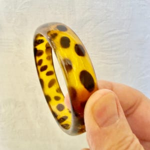 Puede incluir: Un brazalete rígido de color ámbar translúcido que presenta un patrón repetitivo de manchas irregulares de color marrón oscuro a lo largo de toda la banda circular. El accesorio tiene un acabado liso y pulido con un perfil redondeado, evocando la estética clásica del carey.