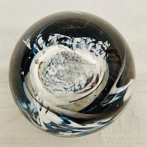 Puede incluir: Un pisapapeles de vidrio redondo con un diseño en espiral. El fondo es negro con detalles en blanco y azul, creando un patrón abstracto dinámico. Se ven pequeñas burbujas de aire dentro del vidrio.