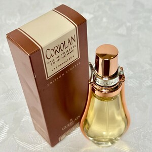 May include: A perfume bottle and box set. The bottle is a clear, teardrop shape with a copper-colored cap and accents. The box is brown with the name "Coriolan" and other text in white. The set is for men.