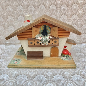 Pode incluir: Uma casa de madeira em estilo chalé com telhado vermelho, paredes brancas e uma porta de madeira marrom. A casa tem uma varanda com uma caixa de flores e um pequeno banco na frente. Há cogumelos vermelhos e plantas verdes ao redor da base da casa.