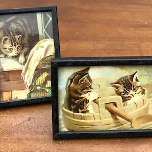May include: Two framed pictures of kittens. The first picture shows a tabby kitten looking out of a window. The second picture shows two tabby kittens in a small wooden boat.