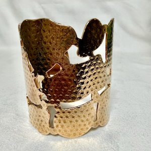 May include: A wide cuff bracelet in a polished rose gold tone metal. The surface features a hammered texture and large, irregular cut-out shapes that create an openwork design. The bracelet has a cylindrical form with a smooth, scalloped edge.