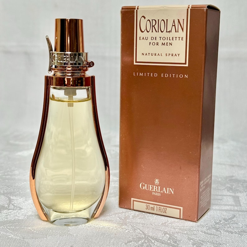 May include: A perfume bottle and box. The bottle is clear with a gold-colored cap and accents, containing a light yellow liquid. The box is brown with the text "CORIOLAN EAU DE TOILETTE FOR MEN NATURAL SPRAY LIMITED EDITION" and "GUERLAIN PARIS".