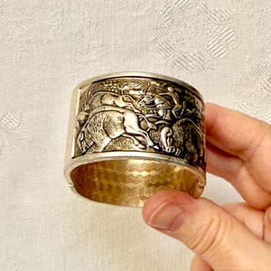 May include: Silver-toned cuff bracelet with a detailed relief depicting a hunting scene with figures, horses, and bison. The bracelet has a textured, gold-colored interior and is held by a hand.