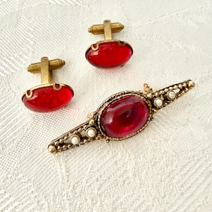May include: A collection of vintage jewellery featuring two gold-toned cufflinks with oval, red glass stones and a gold-toned brooch with a large, oval red stone and small, white pearls. The items are on a white, textured surface.