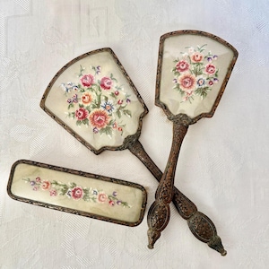 Könnte beinhalten: Ein Toilettenset bestehend aus zwei Handspiegeln und einer rechteckigen Bürste, jeweils mit cremefarbenem Hintergrund und gestickten Blumenmotiven in Rosa, Rot und Blau, eingefasst in verzierte, dunkelmetallische Rahmen mit geprägten Details.
