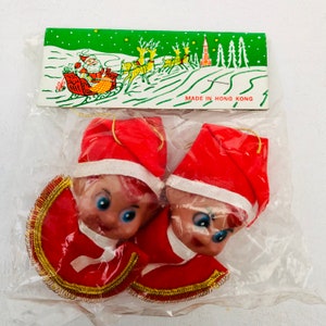 May include: Two vintage Christmas elf ornaments in a clear plastic package. The elves wear red hats and outfits with gold trim. The package includes a green card with a Santa Claus and reindeer scene, and the text "MADE IN HONG KONG".