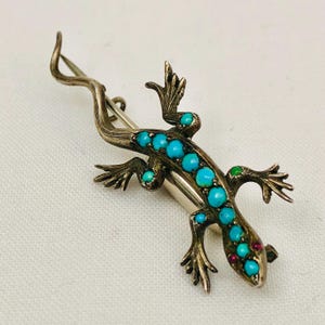 May include: A silver lizard brooch set with turquoise and red gemstones. The lizard is detailed with a textured body and has a long tail.
