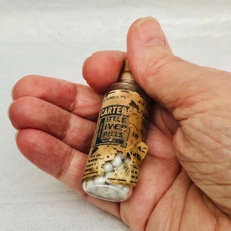 May include: A small glass bottle filled with white pills. The bottle has a label that reads "Carter's Little Liver Pills Registered".
