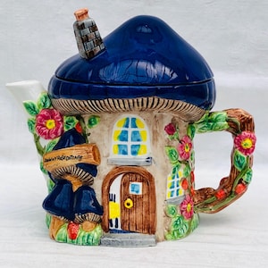 Op de afbeelding: Een grillige keramische theepot in de vorm van een paddenstoelenhuis met een donkerblauw dak en een schoorsteen. De theepot heeft een houten deur, ramen en een handvat versierd met bloemen en bladeren. Het bordje luidt "Downy Rose Cottage".