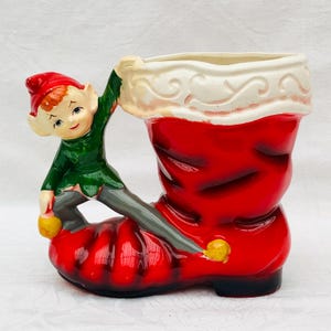 Könnte beinhalten: Ein Keramik-Weihnachtsboot-Pflanztopf mit einem grünen und grauen Elf, der sich am Rand des Stiefels festhält. Der Stiefel ist rot mit weißen Verzierungen und einem weißen Oberteil.