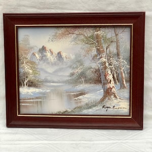 May include: A framed landscape painting of a winter scene with snow-covered trees, a river, and mountains. The painting is set within a dark reddish-brown frame. The artist's signature is visible in the bottom right corner.