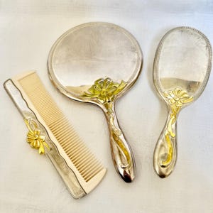 May include: A vanity set consisting of a round hand mirror, an oval hairbrush, and a rectangular comb. Each piece is crafted from silver-toned metal with gold-toned ribbon-like accents. The comb features cream-colored teeth and a metallic handle.