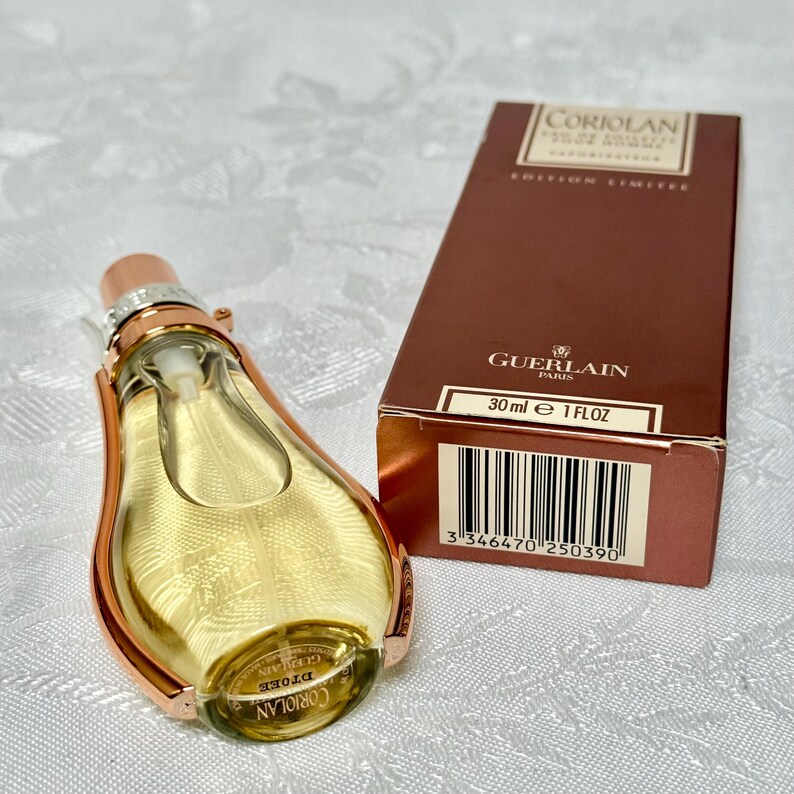 May include: A Guerlain Coriolan perfume bottle with a copper-colored frame and a gold-colored liquid. The bottle is next to a brown box with the text "Coriolan" and "Guerlain Paris". The box indicates the perfume is 1 fl oz.