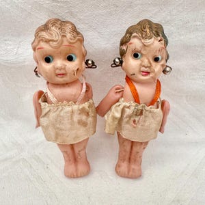 May include: Two vintage bisque dolls with painted facial features, blue eyes, and curly hair. They wear small, cream-colored skirts and have tiny, attached wings. Each doll has a small earring. One doll has an orange necklace.