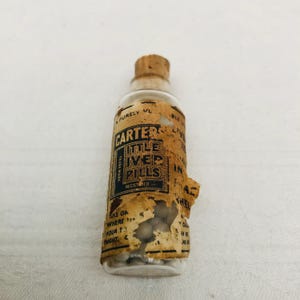 May include: A small glass bottle with a cork stopper filled with white pills. The label reads "Carter's Little Liver Pills, Trademark Registered".