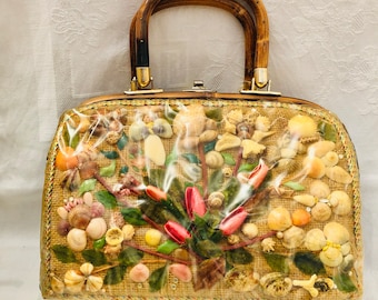 Könnte beinhalten: Eine Vintage-Handtasche mit Bambusgriff und klarer Kunststoffauflage. Die Tasche ist mit Muscheln, Kunstblumen und Grünzeug verziert. Die Tasche hat eine rechteckige Form mit abgerundeten Kanten und goldfarbener Hardware.
