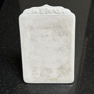 May include: A white ceramic recipe holder with a decorative top edge. The rectangular holder has a slightly textured surface with handwritten recipe notes in pencil. The word "MENU" is at the top.
