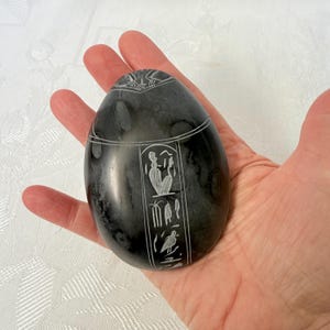 Peut inclure: Une sculpture en pierre sombre, de forme ovoïde, avec des hiéroglyphes et des figures gravés en blanc. La pierre polie est tenue dans une main, mettant en valeur les détails complexes du design d'inspiration égyptienne antique. La sculpture présente un oiseau et d'autres symboles.