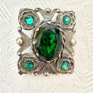 May include: Vintage silver-toned brooch with a large, oval, emerald-green gemstone at the center. The brooch features smaller, round, green gemstones and floral accents. The design is ornate and detailed, with a rectangular shape.