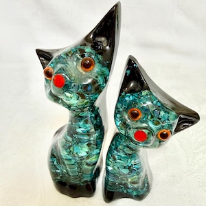 May include: Two cat figurines crafted from glass with a black finish on the ears and base. The bodies feature a mosaic of teal and blue glass fragments, accented by bright orange circular eyes and a small, vibrant red circular nose on each face.