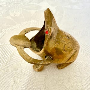 Pode incluir: Um cinzeiro vintage em forma de rã de latão com boca aberta e olho de gema vermelha. O cinzeiro tem uma pega e é decorado com detalhes gravados. A rã está colocada numa superfície branca.