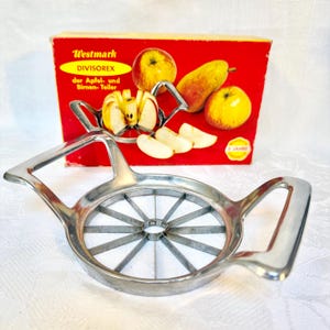 Könnte beinhalten: Ein Vintage Westmark Divisorex Apfel- und Birnenschneider. Der Schneider ist aus glänzendem Metall gefertigt und hat ein kreisförmiges Klingendesign mit Griffen. Die rote Schachtel dahinter zeigt Äpfel und Birnen und den Text "Westmark Divisorex".
