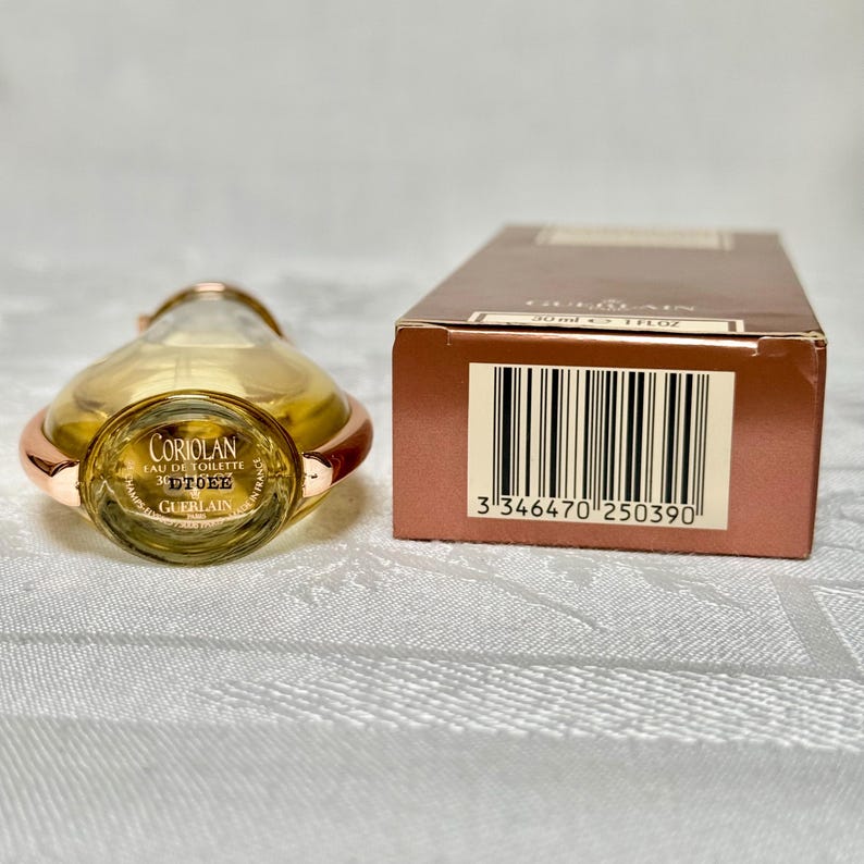May include: A Guerlain Coriolan Eau de Toilette perfume bottle with a gold-colored top and accents. The bottle is next to a brown box with a barcode. The bottle has the text "Coriolan Eau de Toilette" and "Guerlain" on it.