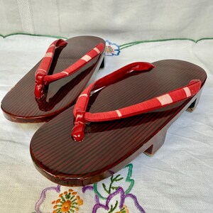 Könnte beinhalten: Ein Paar traditionelle japanische Geta-Sandalen. Die Holzbasis ist dunkelrot mit einem Streifenmuster, und die Riemen sind rot mit weißen Streifen. Die Sandalen haben einen kleinen Absatz und sind für den Außenbereich konzipiert.