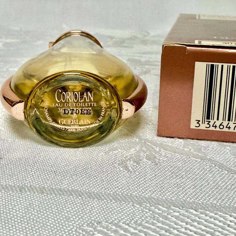 May include: A bottle of Guerlain Coriolan Eau de Toilette with a gold-colored bottle and rose gold accents. The bottle is lying on its side, with the label visible. A brown box is partially visible in the background.