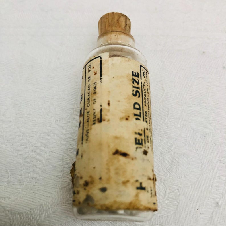 May include: A small, clear glass bottle with a cork stopper and a label that reads "ELD SIZE" and "AFTER PRODUCTS INC".