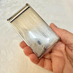 May include: A rectangular, silver-toned cigarette case with a hinged lid. The case features an etched, linear pattern across its surface. The case is held in a hand, showcasing its size and design.