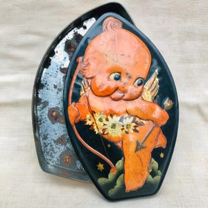 May include: A vintage tin box with a black background and a colorful image of a cherub holding a bow and arrow. The cherub is orange with a yellow halo and a white sash. The box is decorated with gold stars and clouds.