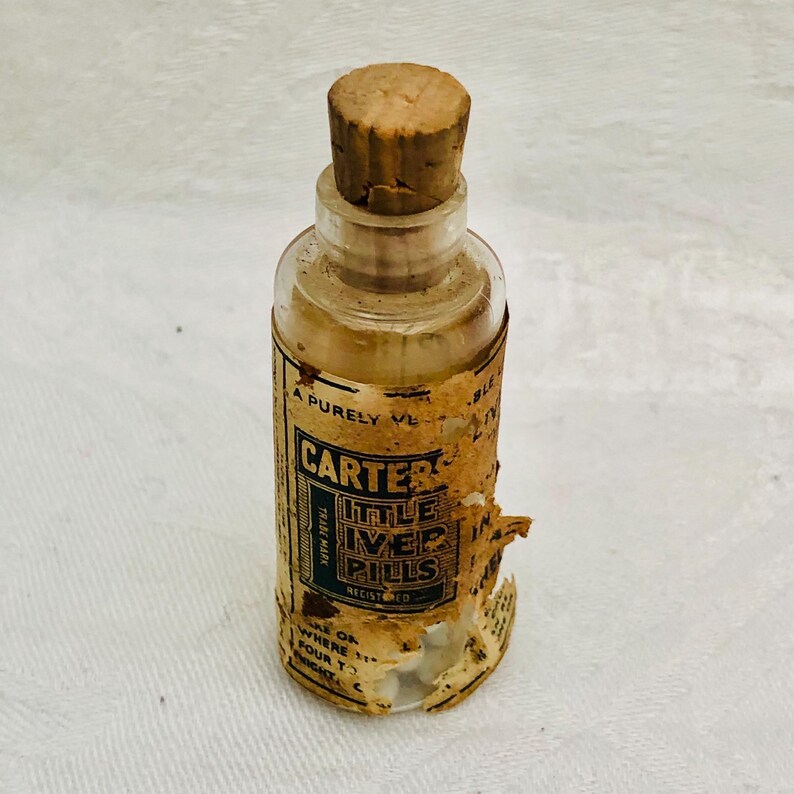 May include: A vintage glass bottle with a cork stopper. The bottle is labeled "Carter's Little Liver Pills" and has a green and white design. The bottle is filled with white pills.